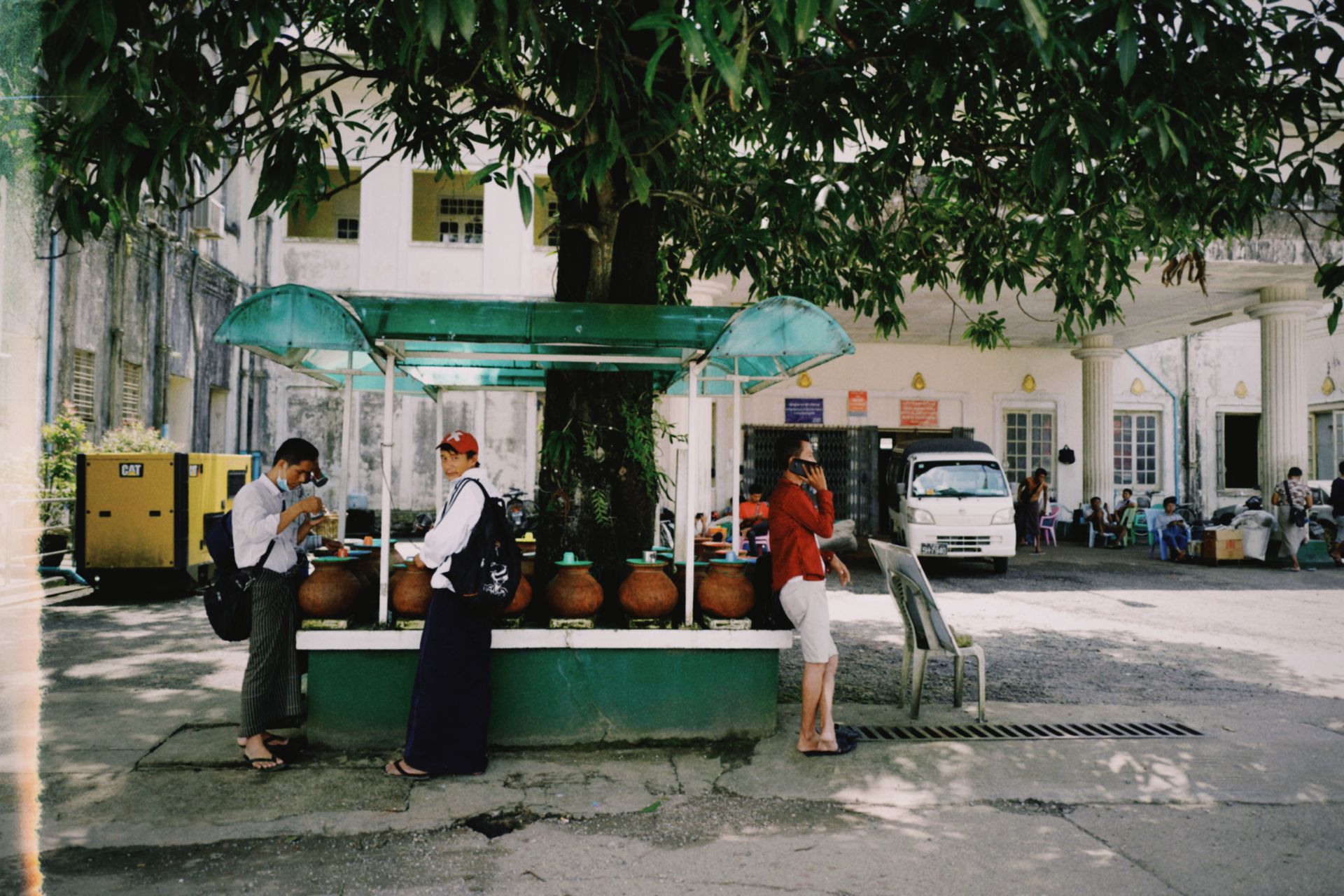 Urban street scene with three people interacting around clay pots under a tree.