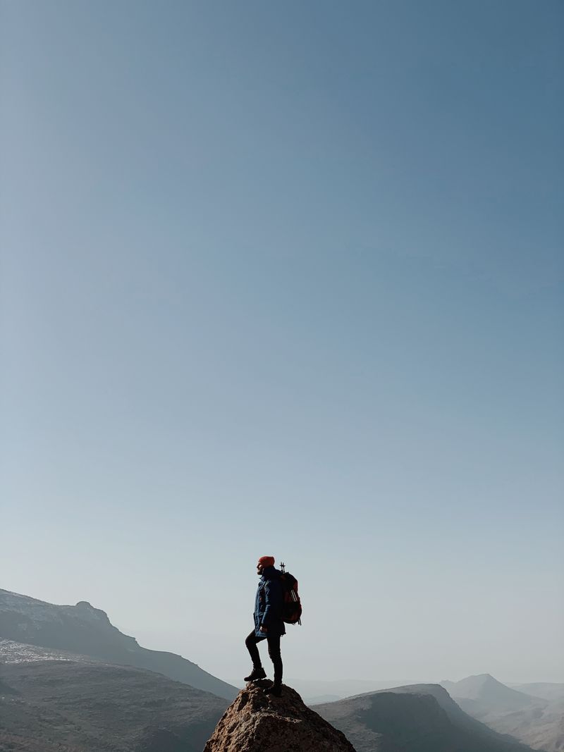 A lone hiker stands triumphantly on a mountain peak under a vast blue sky