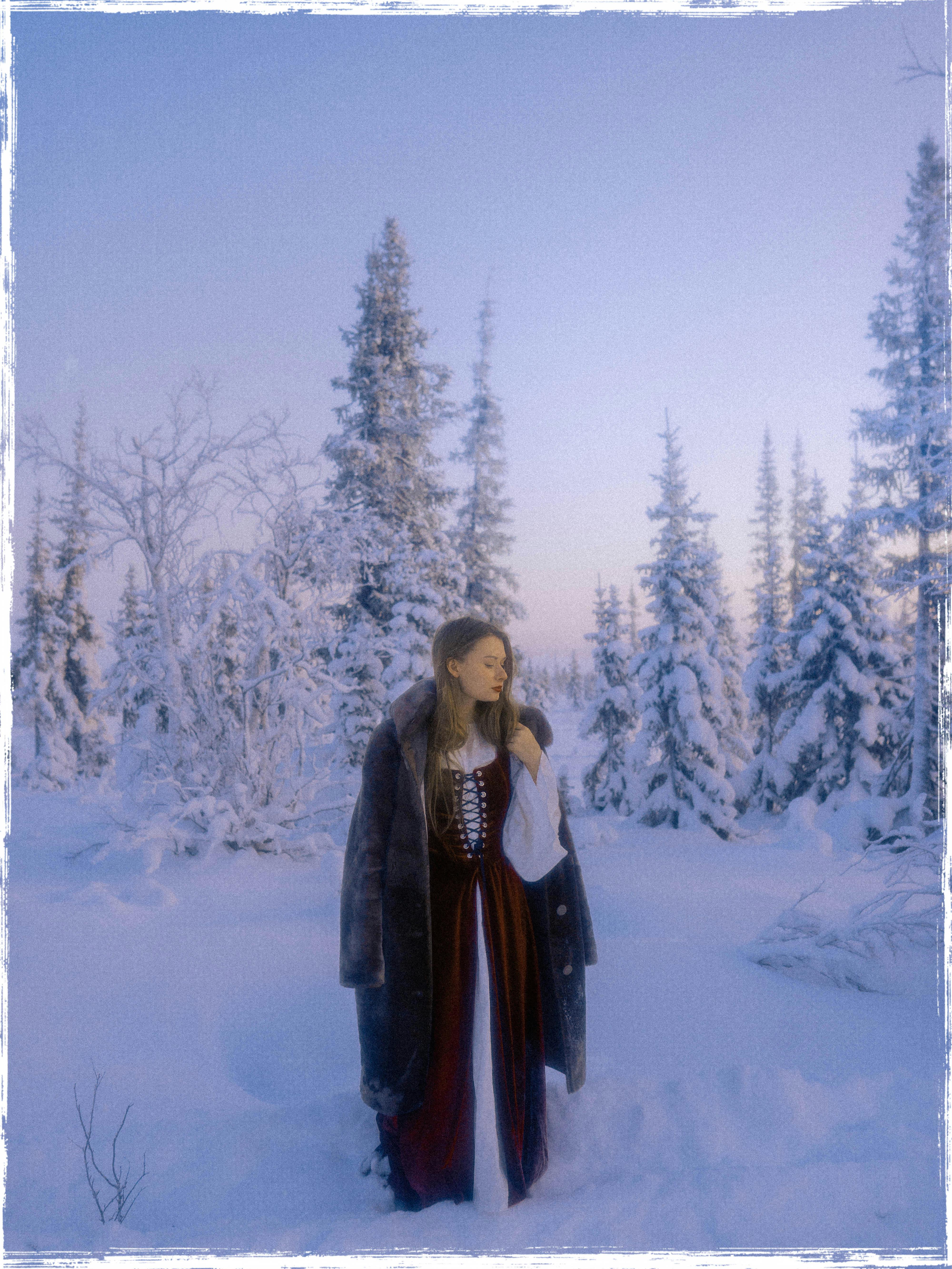 A woman in traditional attire stands in a snow-covered forest, evoking winter serenity.