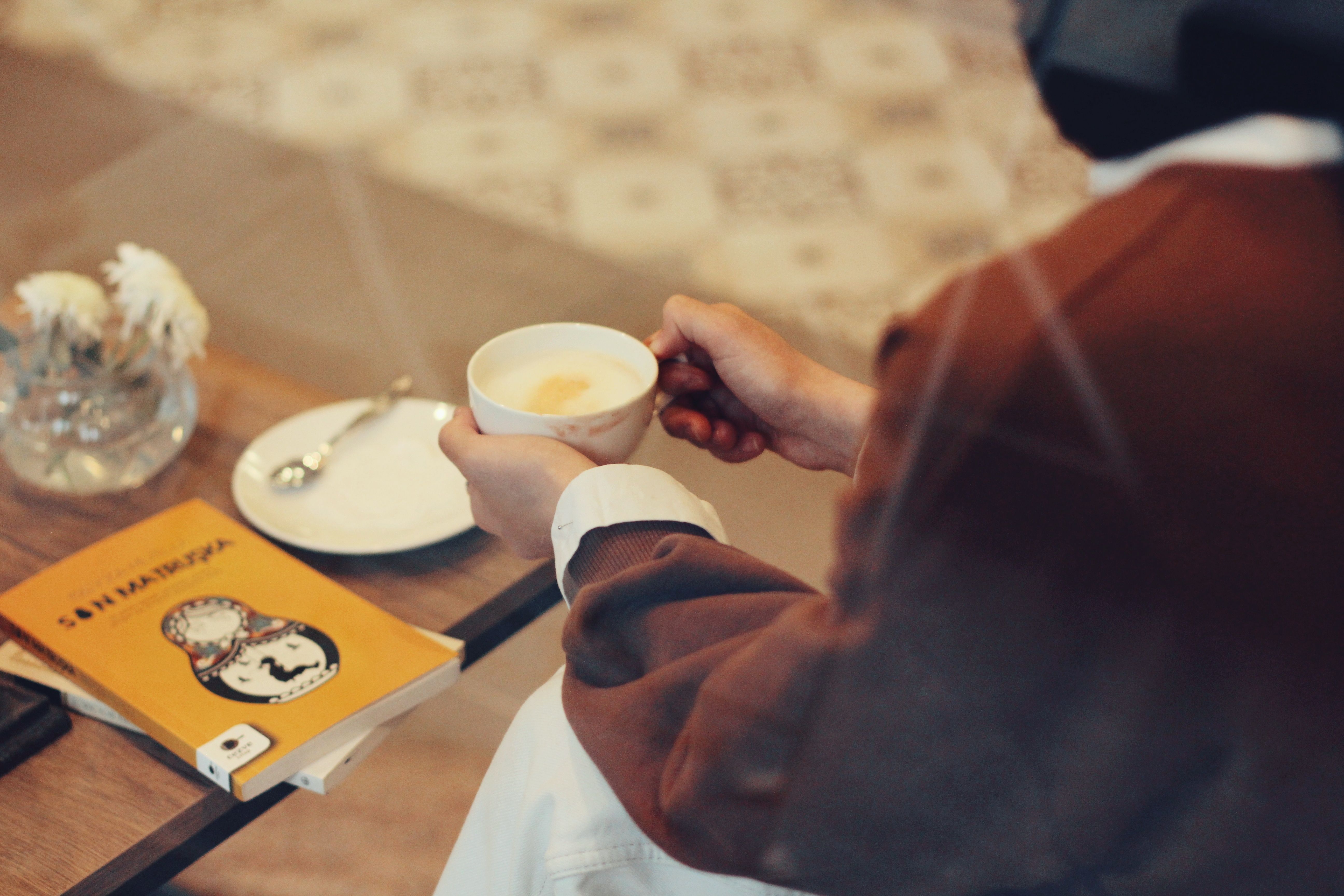 Warm scene of an individual holding a coffee cup in a cafe while reading a book on a wooden table.