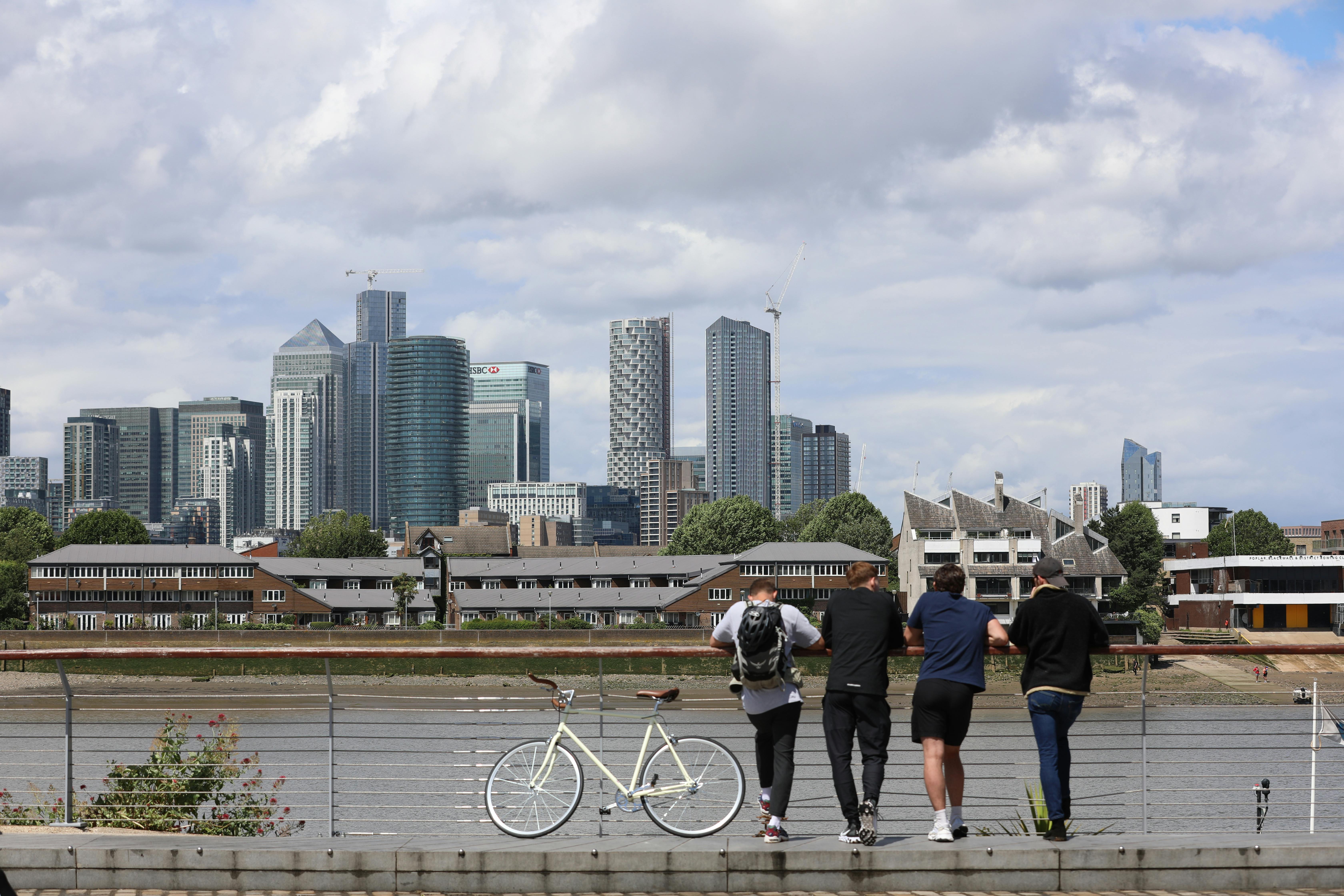 Group of young adults relax by the river with bicycles, overlooking London's modern skyline.