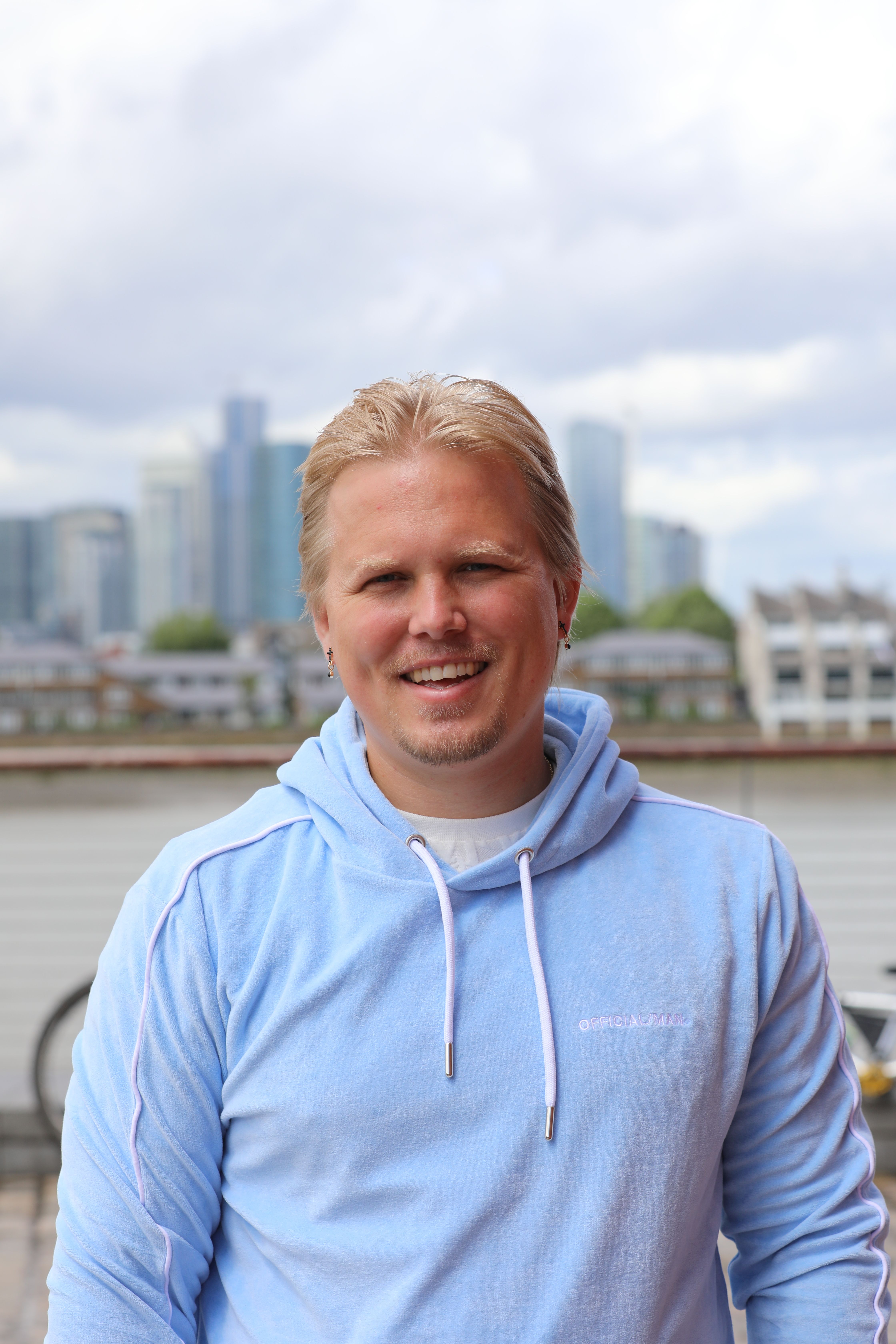 Man smiling in a blue hoodie with a cloudy city skyline in London.