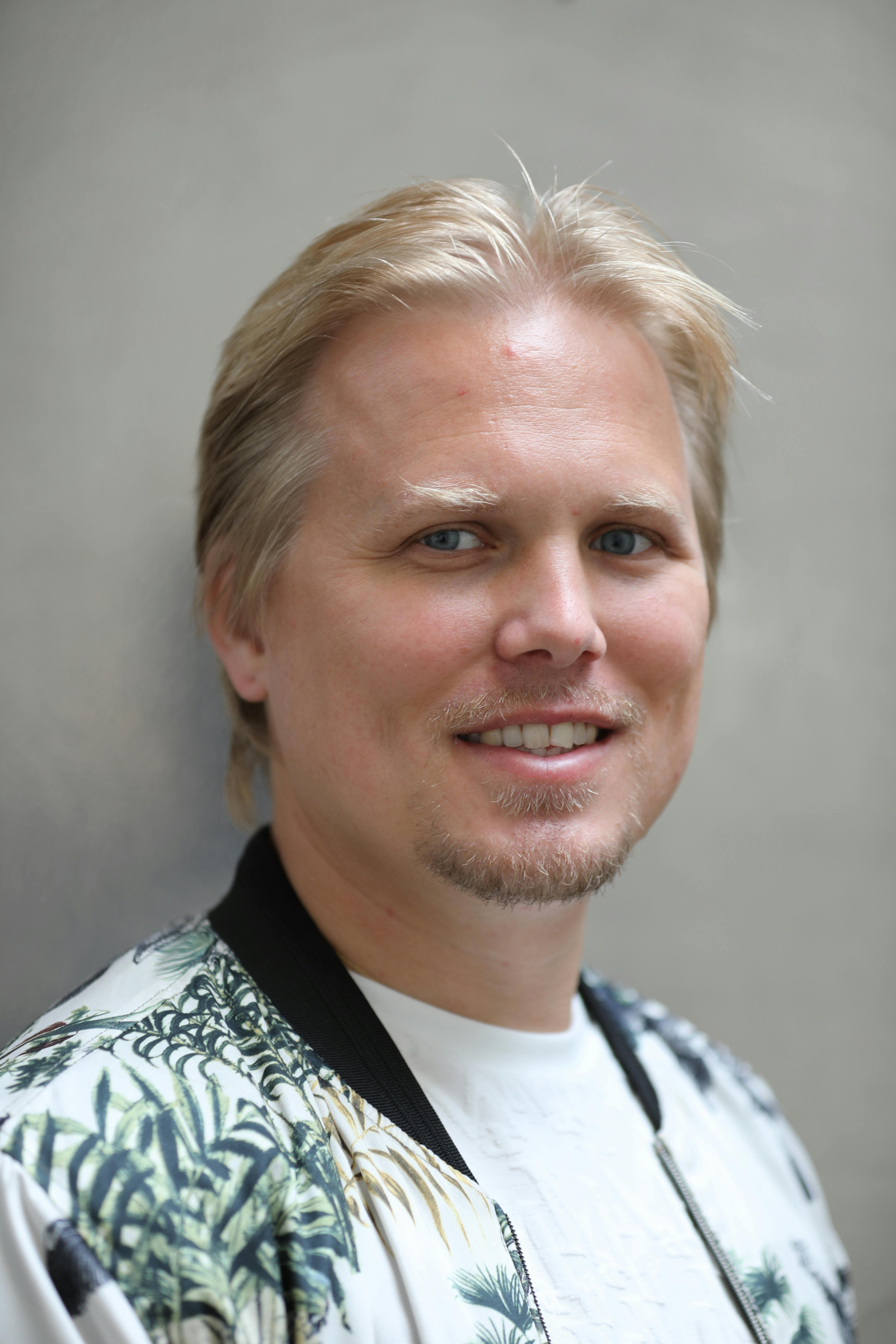 A smiling man with blond hair wearing a floral shirt in an urban setting.