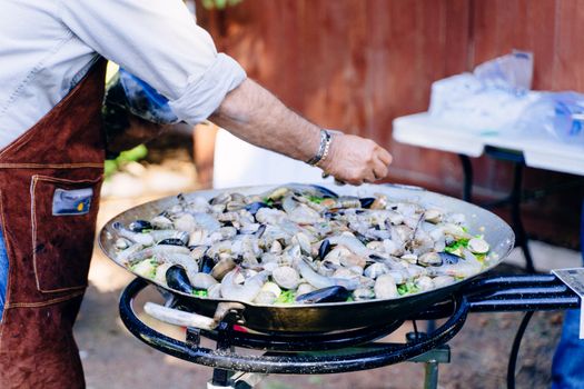A man in an apron cooking a seafood dish with clams and mussels in a large pan.