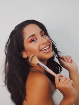 Smiling woman with colorful makeup and sequins, holding a brush in a studio setting.