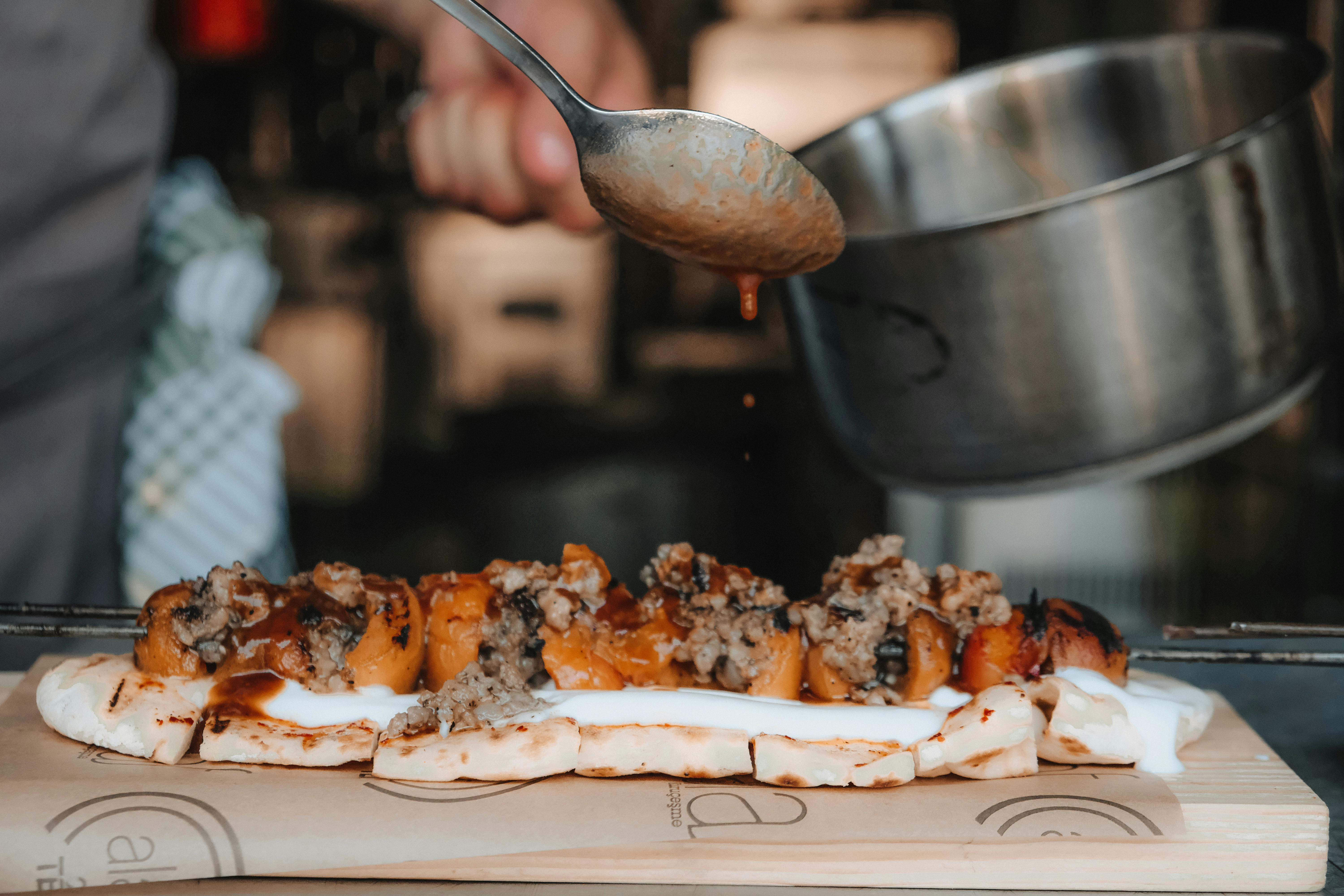 Close-up of a chef pouring sauce on grilled flatbread with meat and veggies, highlighting delicious preparation.