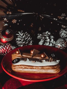 Delicious eclair on a red plate with festive pine cones and ornaments.