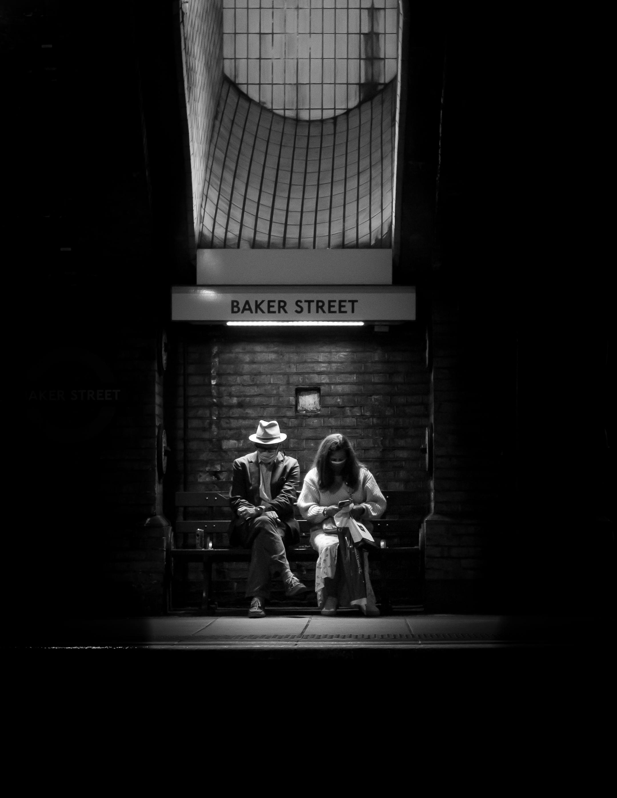 Moody black and white photo of two people sitting at Baker Street station, London.