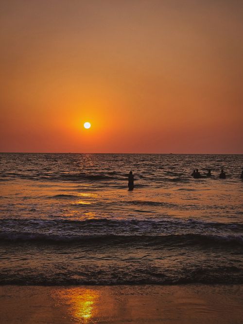 Free Silhouetted people enjoy a serene sunset on a tranquil beach with gentle waves and a warm golden sky. Stock Photo