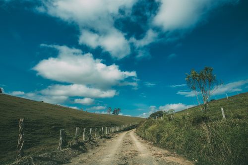 Free Beautiful rural dirt road winding through a green countryside landscape with a vivid blue sky and scattered clouds. Stock Photo