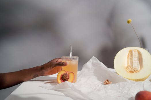 A refreshing glass of melon and peach juice being held by hand with fresh fruits and paper background.