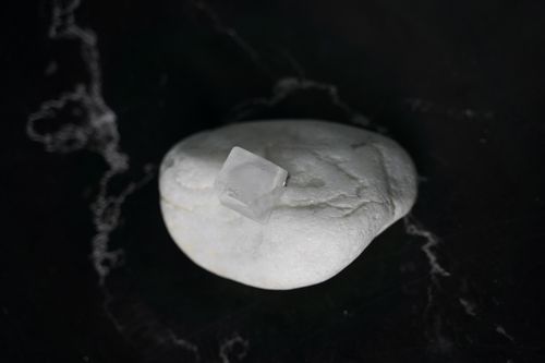 Free A minimalist still life of an ice cube resting on a smooth white stone against a black marble background. Stock Photo