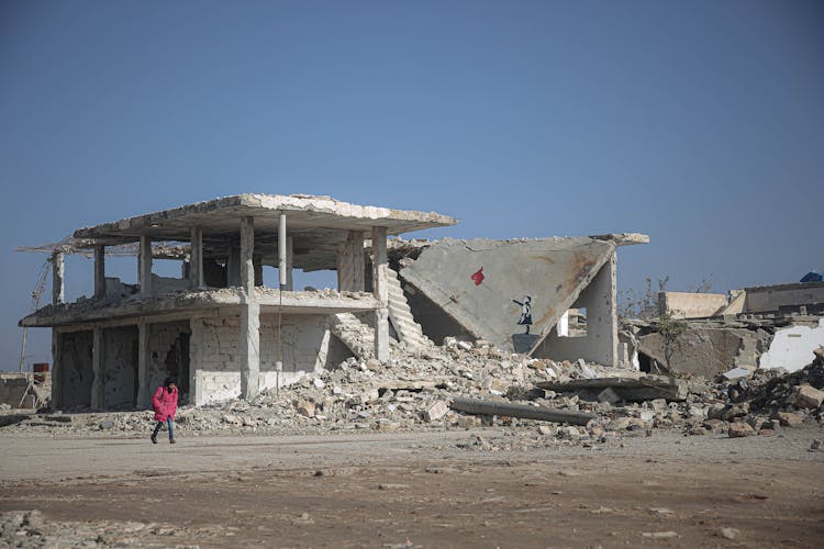 A Child Wearing A Pink Coat Walking Past A Broken Building