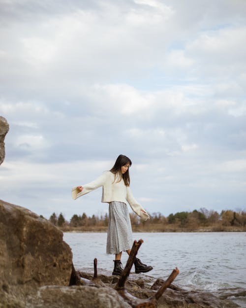 Woman in White Sweater Walking on Rocks Beside a River