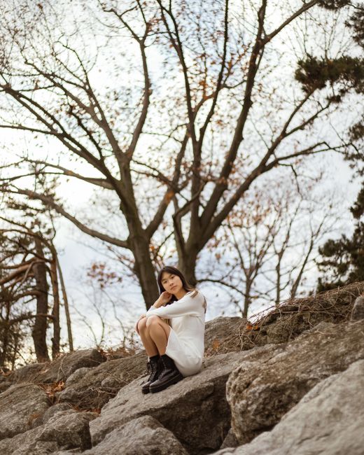 A woman sits on rocks in an outdoor setting with bare trees, creating a serene and contemplative mood.
