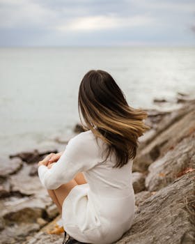 A woman in a white dress with dyed hair sitting on rocks by the sea.