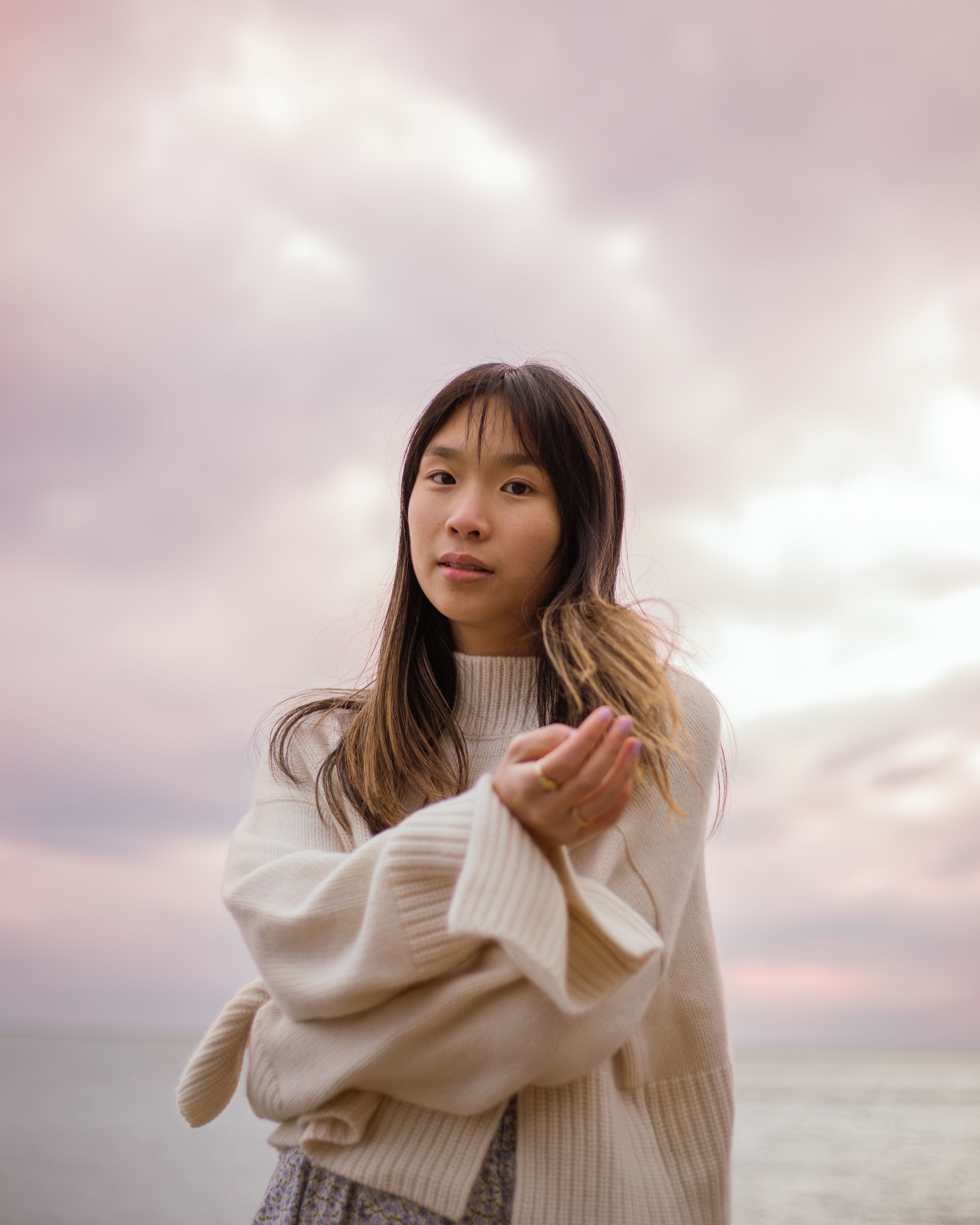 Asian woman in a cozy turtleneck sweater standing by the ocean during a pastel sunset, exuding tranquility.