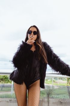 Fashionable adult woman in fur coat and sunglasses smoking outdoors.