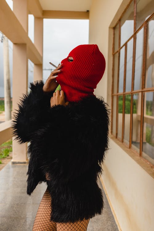 Person in Black Fur Coat and Red Knit Cap Covering Face With Black Fur Scarf