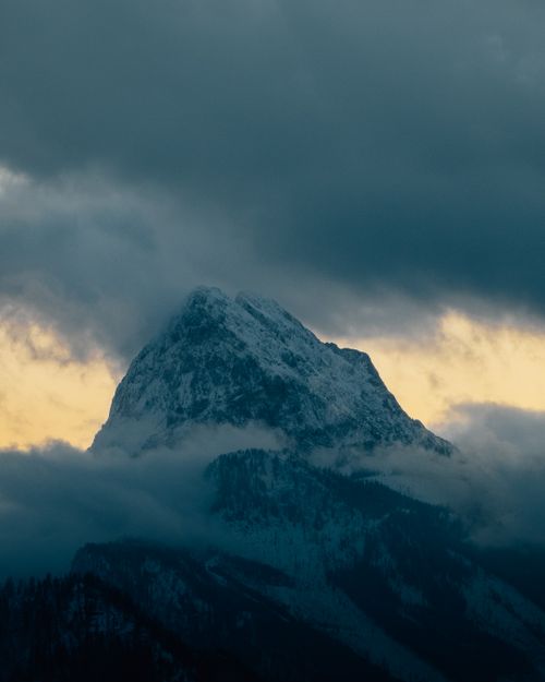 Free A breathtaking view of a snow-covered mountain peak surrounded by clouds at dusk. Perfect for winter-themed projects. Stock Photo