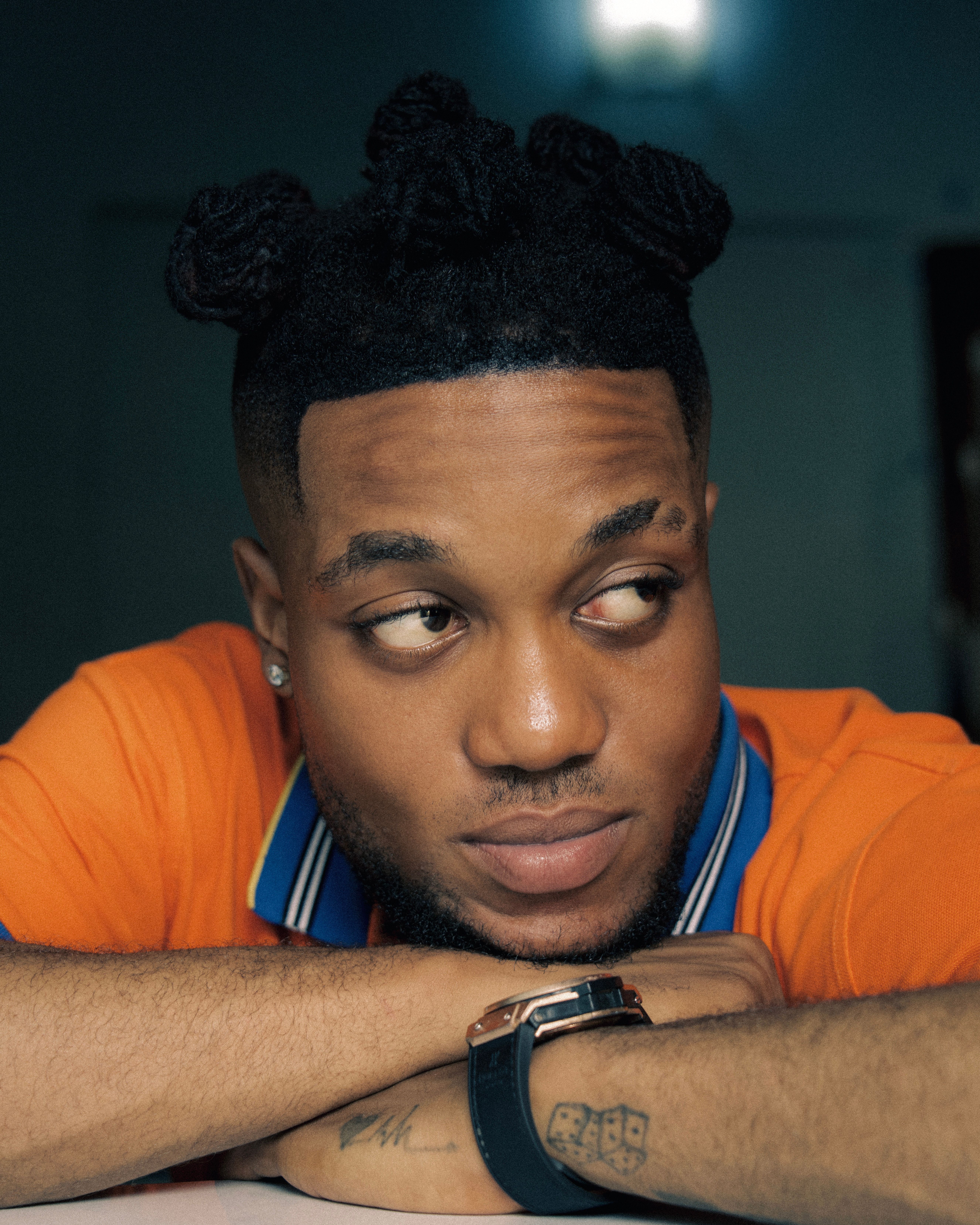 Close-up portrait of a man with tattoos, resting his chin on hands, wearing an orange shirt.