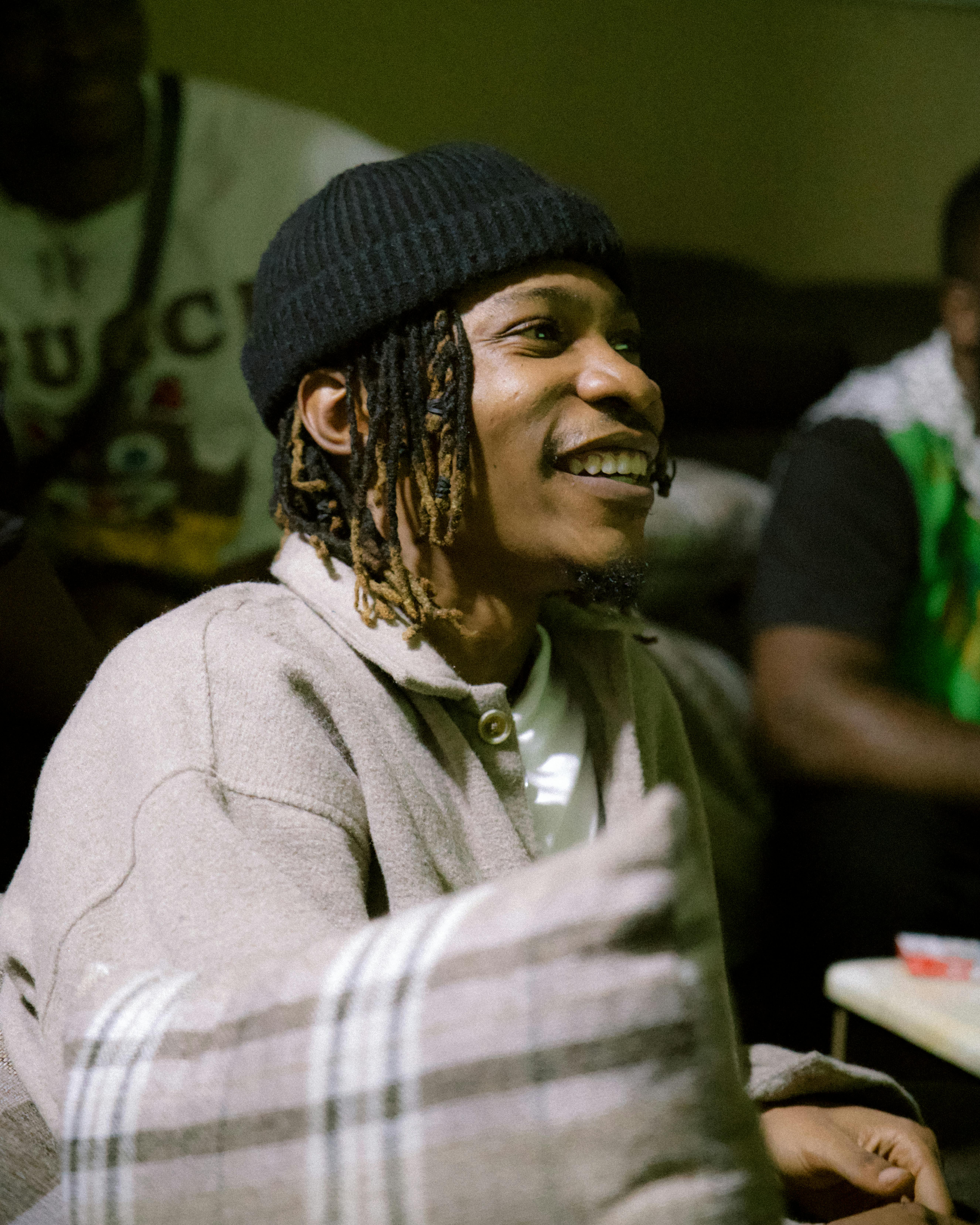 African American man with dreadlocks smiling warmly indoors, wearing a beanie and gray sweater.