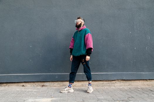 Stylish man wearing colorful sweater and jeans stands by gray wall.