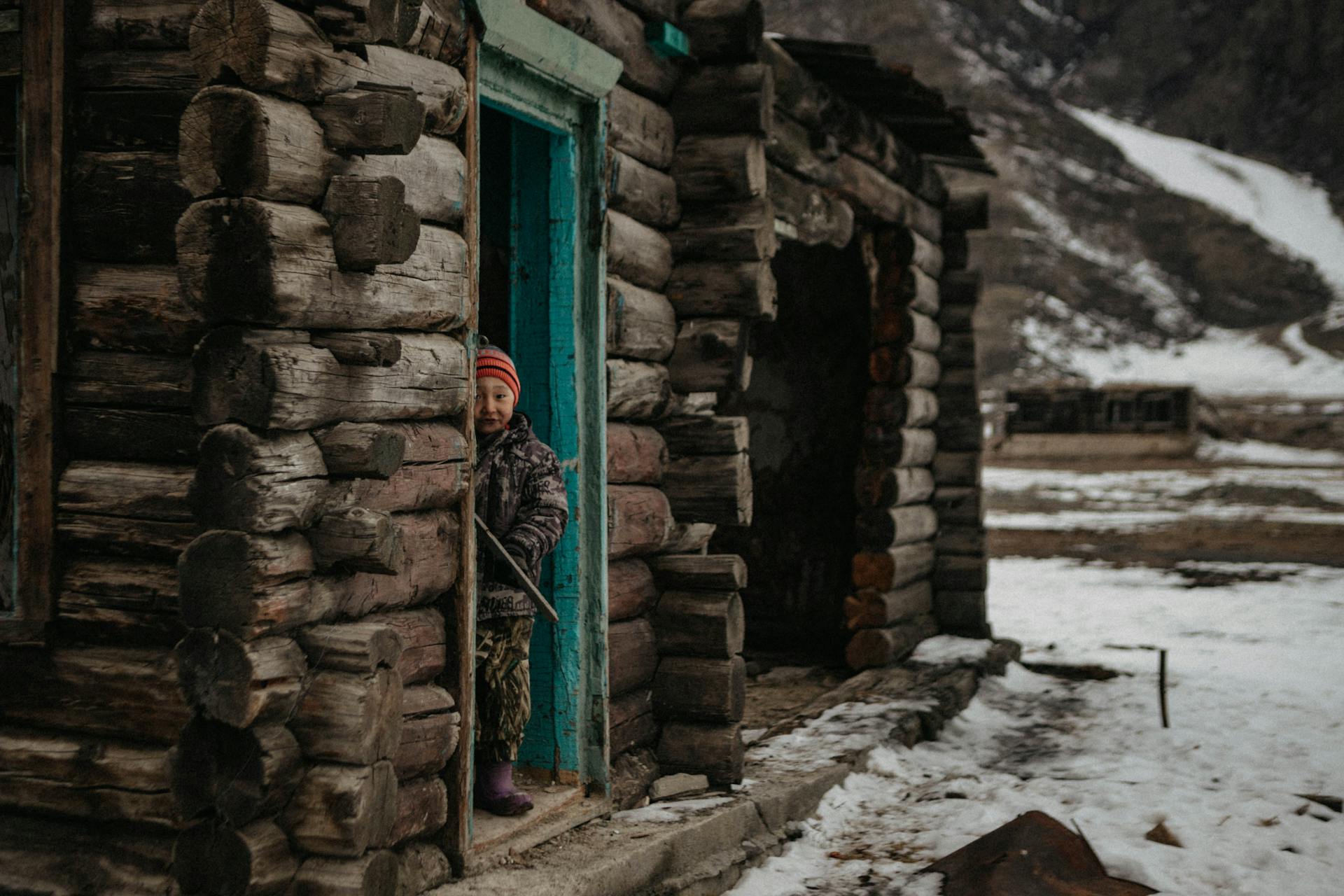 Young child peeks from a rustic log cabin door amidst snowy Altai landscape.