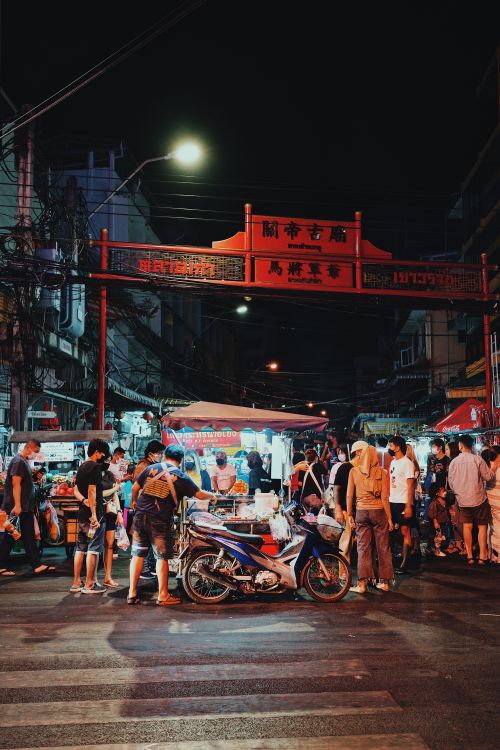 Free Vibrant night market in a city with people shopping and street food vendors near a traditional archway. Stock Photo