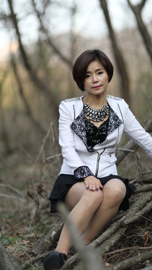 Woman in White Coat Sitting on the Brach of the Trees on the Ground while Looking at the Camera