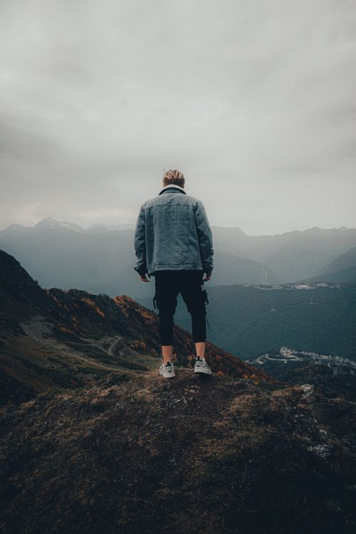 Free Man in denim jacket standing on mountain, gazing at moody sky and distant mountains, creating a sense of solitude. Stock Photo