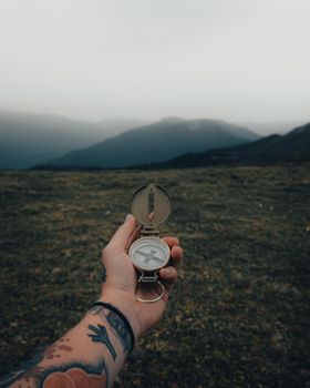 A tattooed hand holding a compass against a backdrop of foggy mountains, showcasing exploration and adventure.