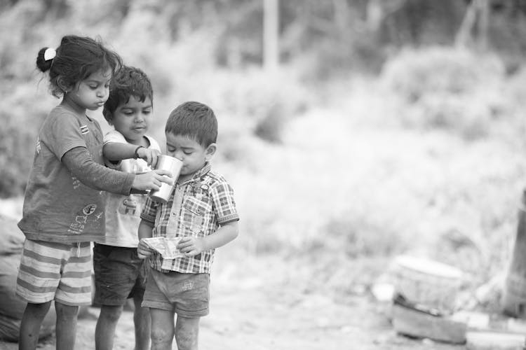 Grayscale Photo Of Kids Helping A Child Drink From A Cup