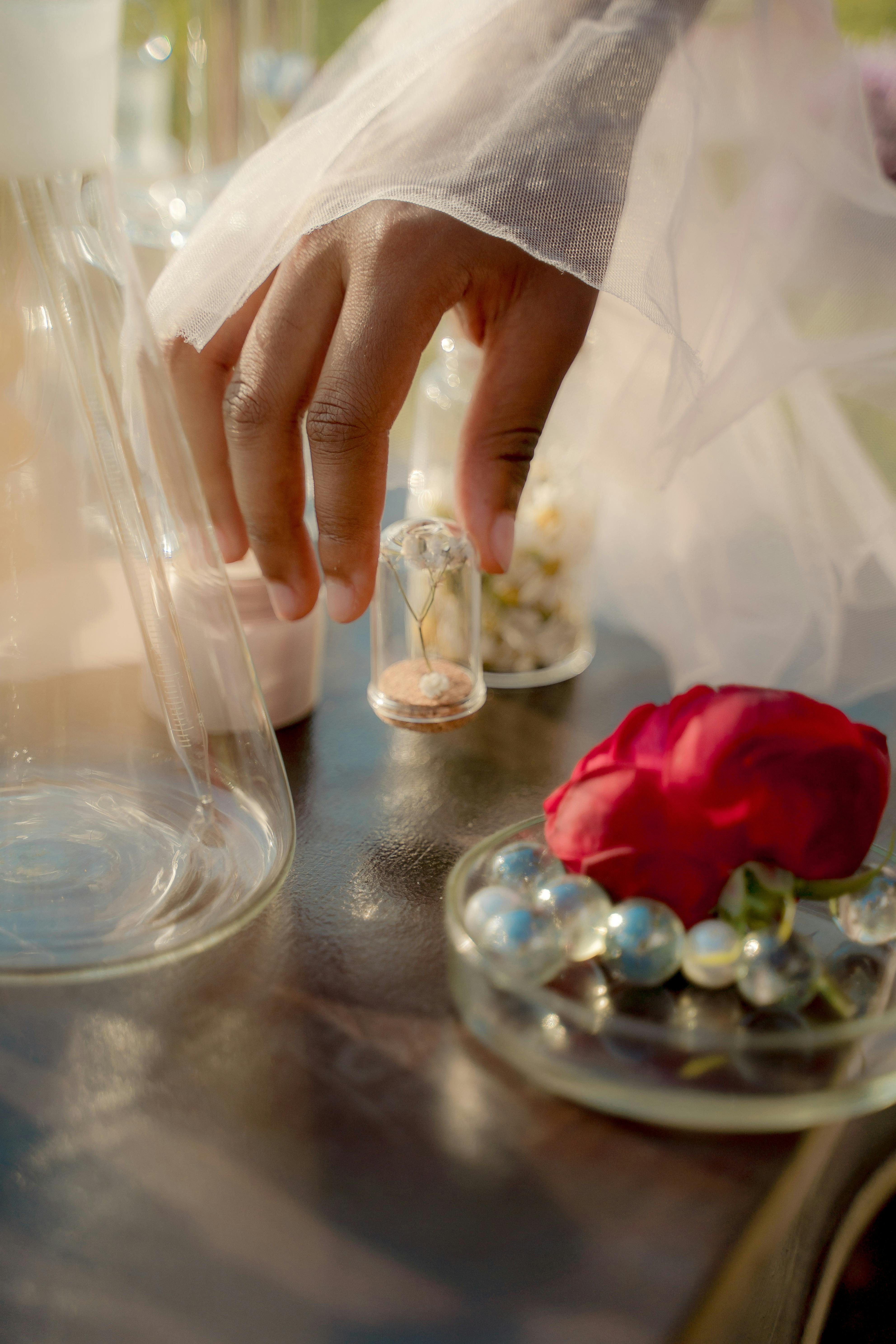 A gently touching hand holding a small glass vial near a rose and pearls, creating a dreamy composition.