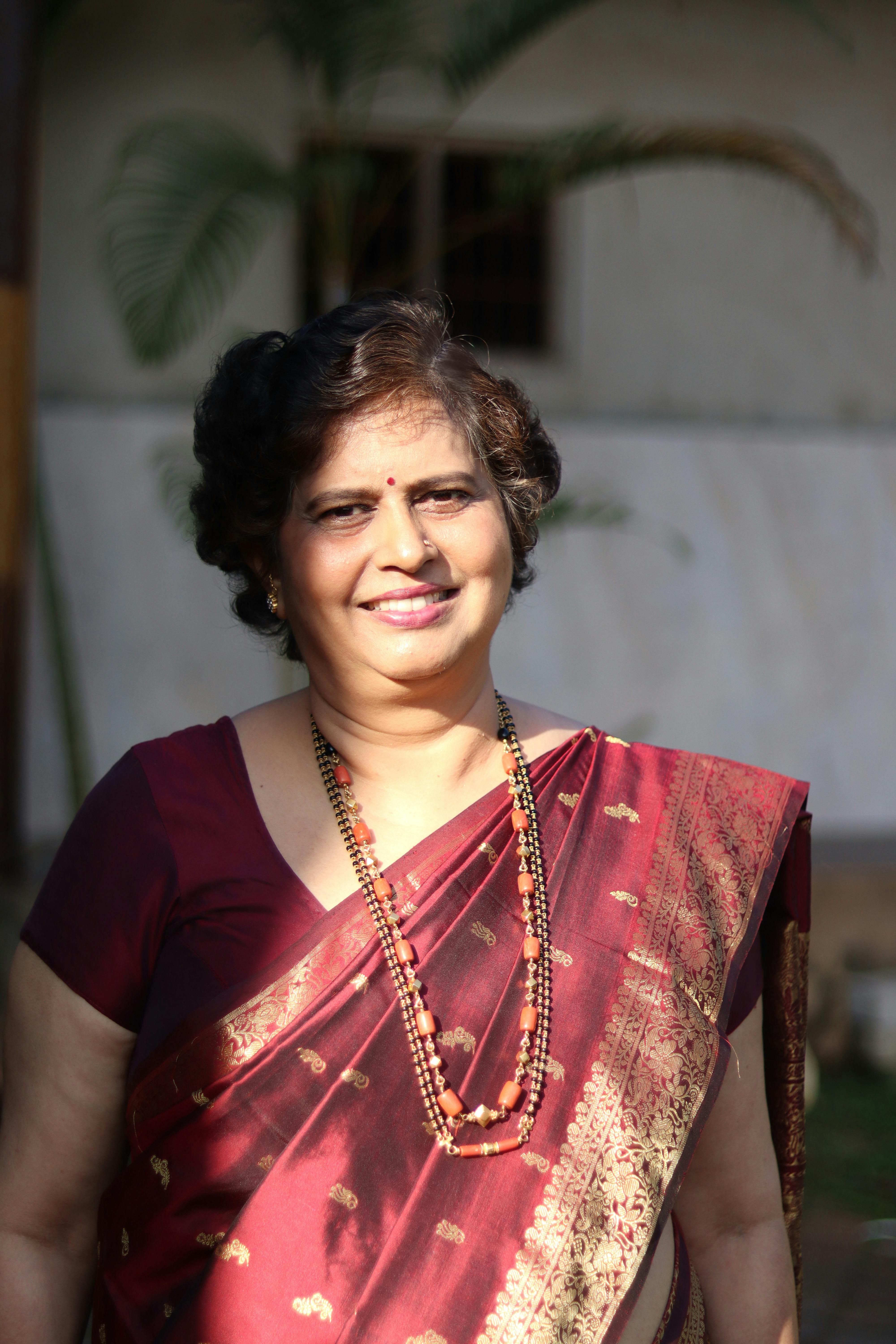 South Asian woman smiling outdoors in a maroon sari, adorned with necklaces.