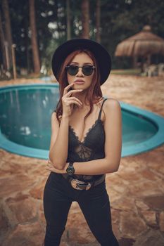 Chic woman in black lace top, sunglasses and hat posing confidently by a pool outdoors.