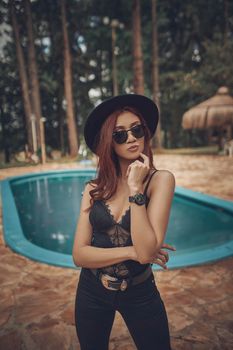 Stylish woman in sunglasses and chic outfit posing at a poolside with a forest backdrop in Paraguay.