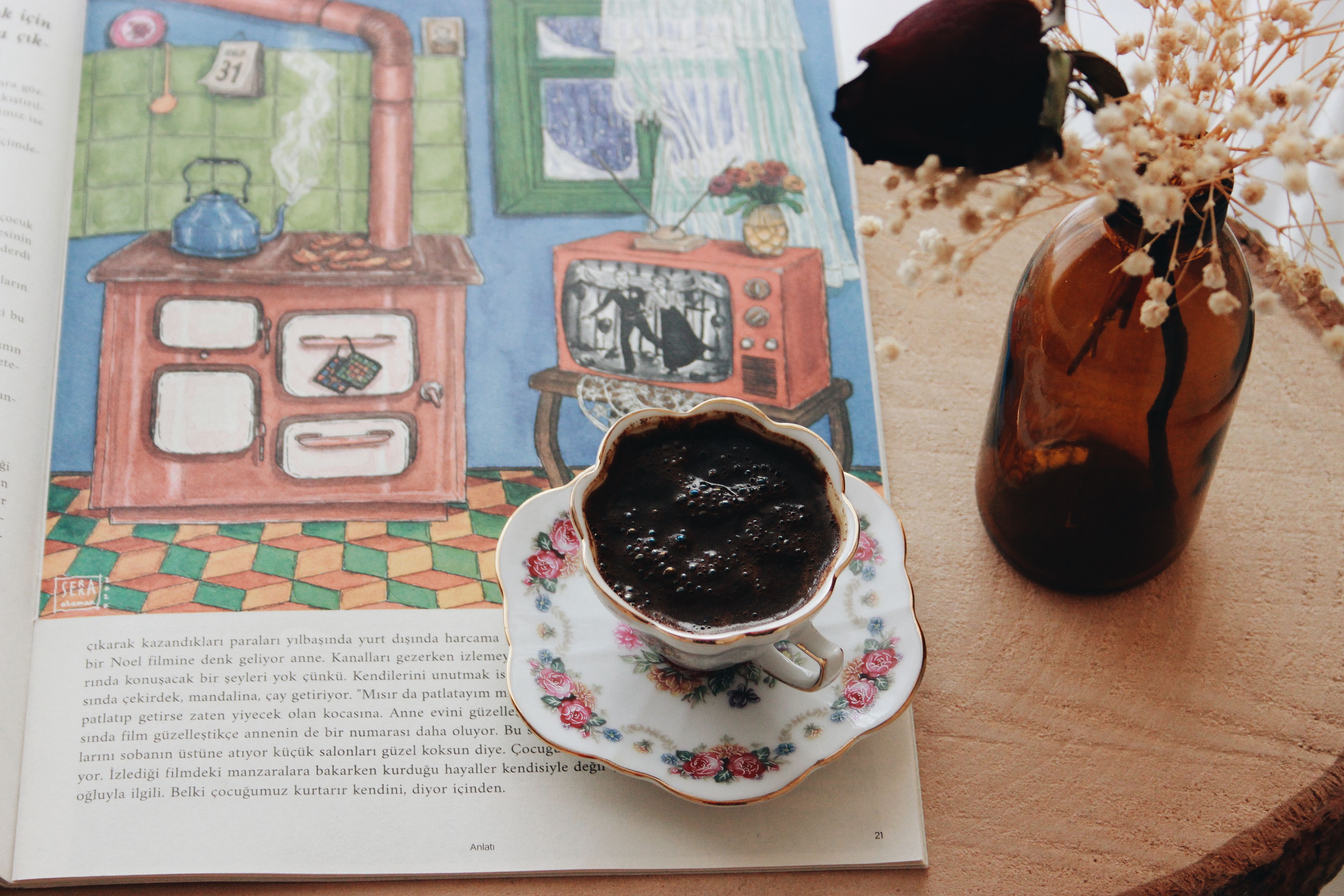 A cozy scene featuring Turkish coffee, floral decor, a book, and a rustic glass bottle on a wooden surface.