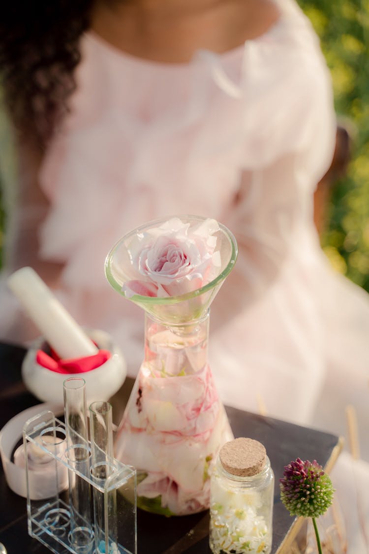 Unrecognizable Woman Sitting At Table With Glass Flask Full Of Rose Petals