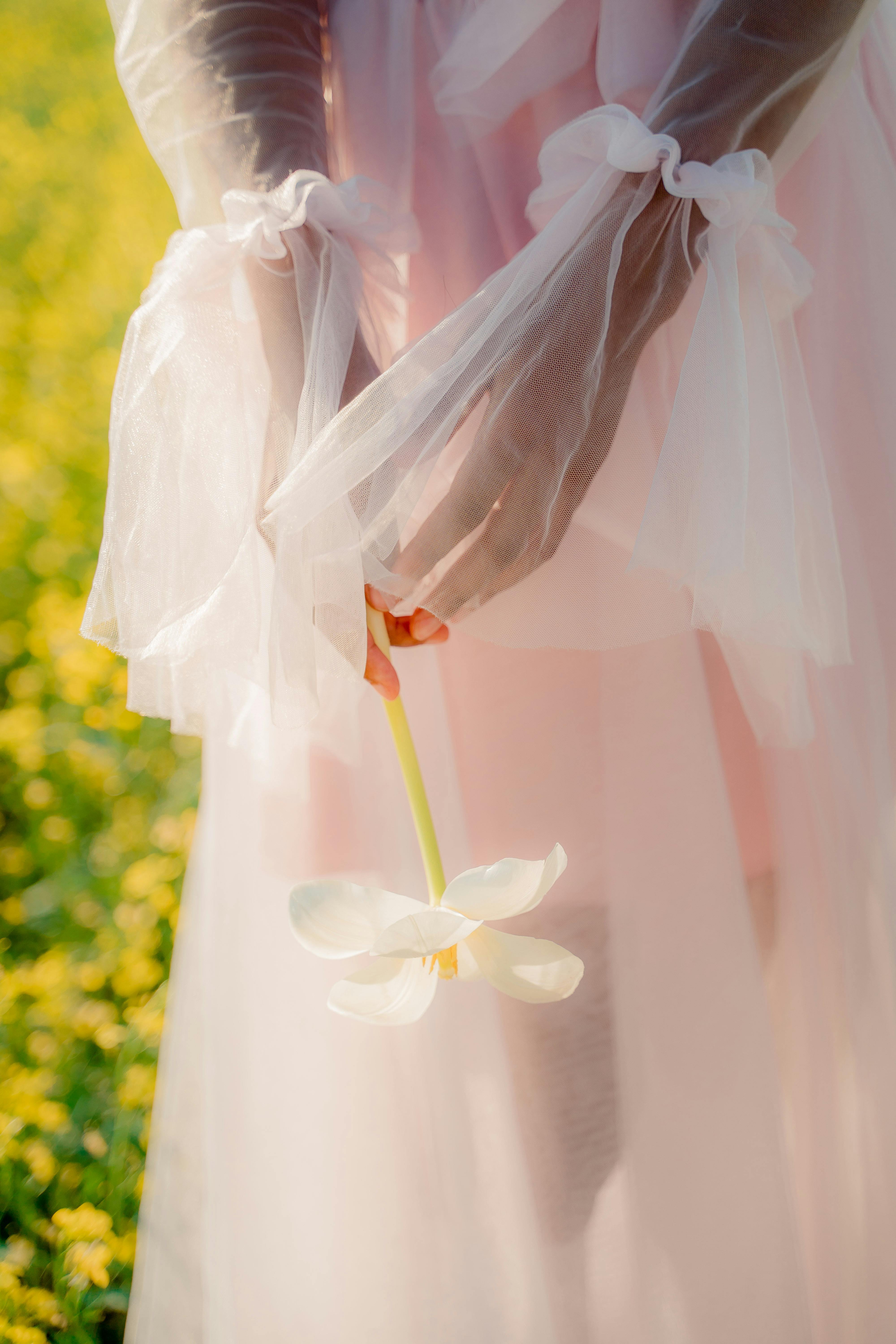 A delicate woman in a pink dress gracefully holding a white flower in a sunny meadow.