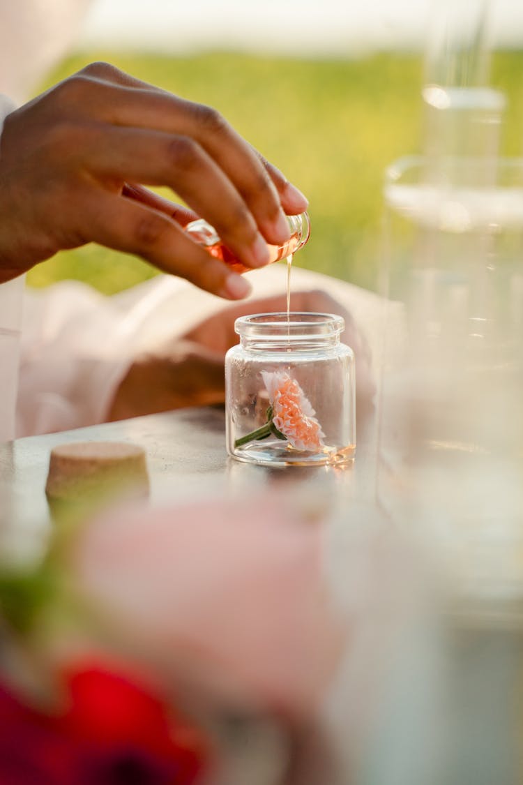 Unrecognizable Female Hand Pouring Oil Onto Flower In Glass Jar