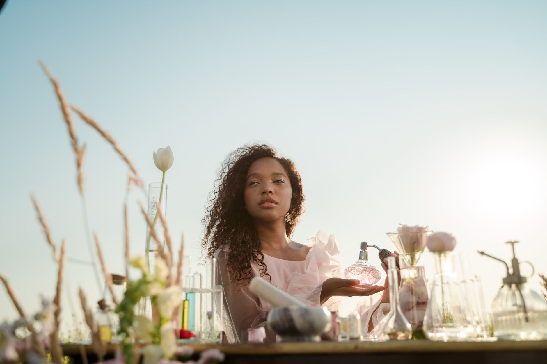 A woman with curly hair holding a glass bottle in a floral and laboratory-themed outdoor setting.