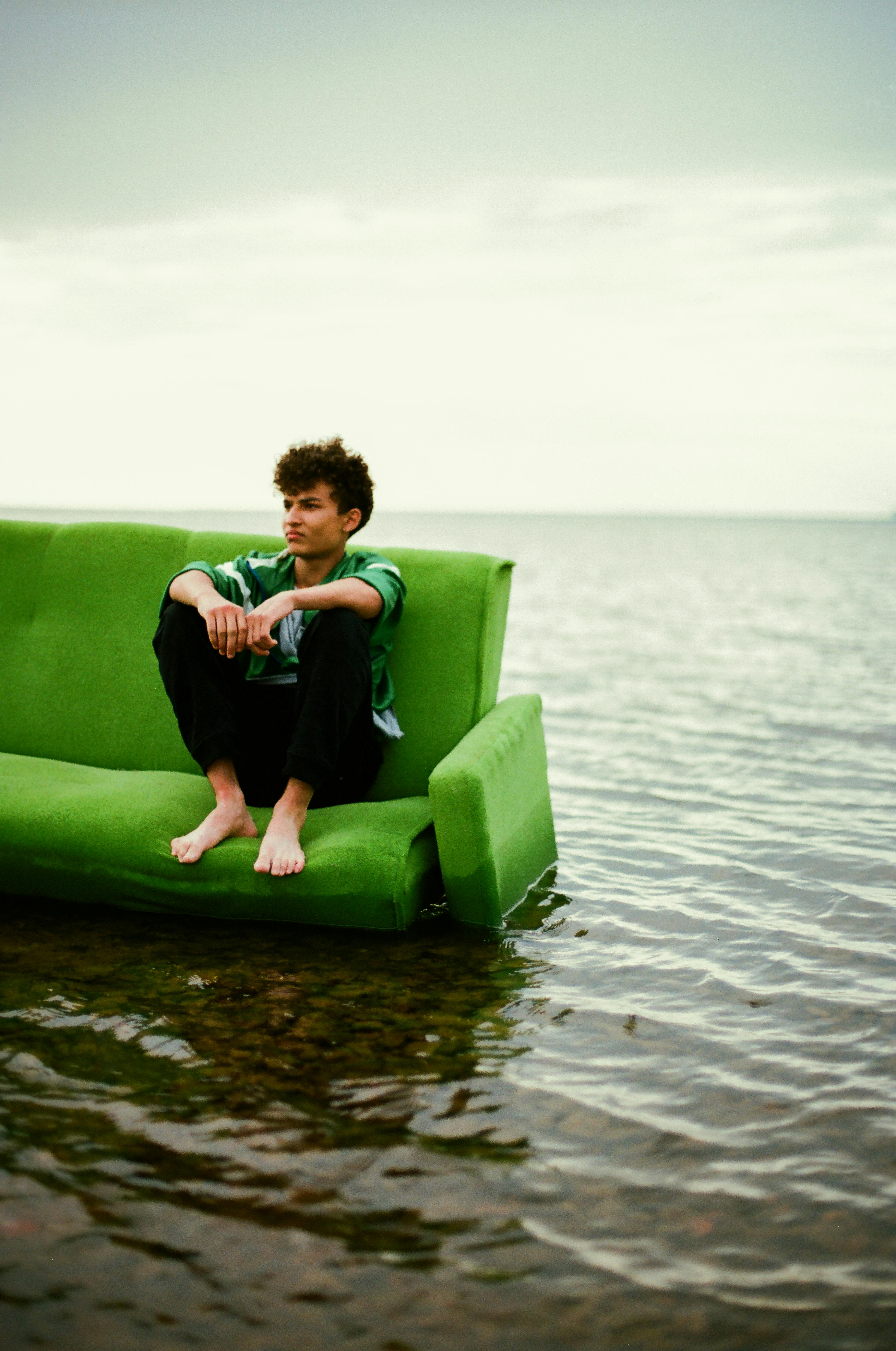 A young man sits contemplatively on a green sofa placed in shallow river water.