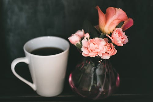 コーヒーのマグカップの横にある透明なガラスの花瓶にピンクのカーネーションの花とピンクのバラの花