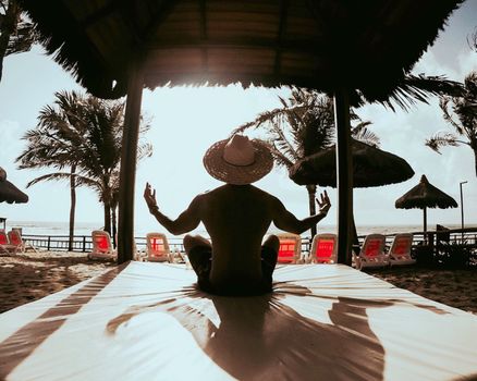 A man meditates in the lotus position on a tropical beach under a nipa hut, embracing summer relaxation.