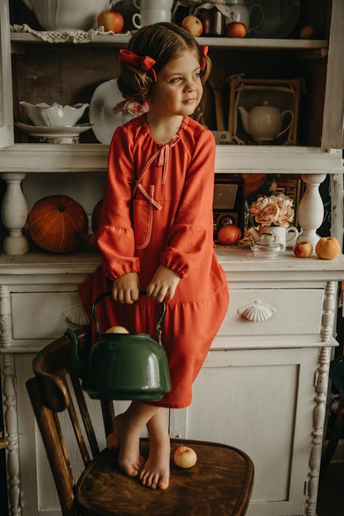 Girl in Red Dress Standing on Chair Holding Kettle