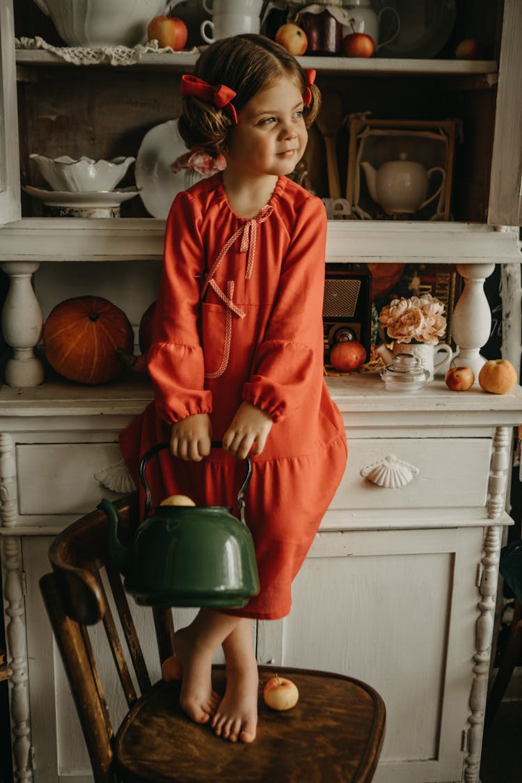 Girl In Red Dress Standing On Chair Holding Kettle