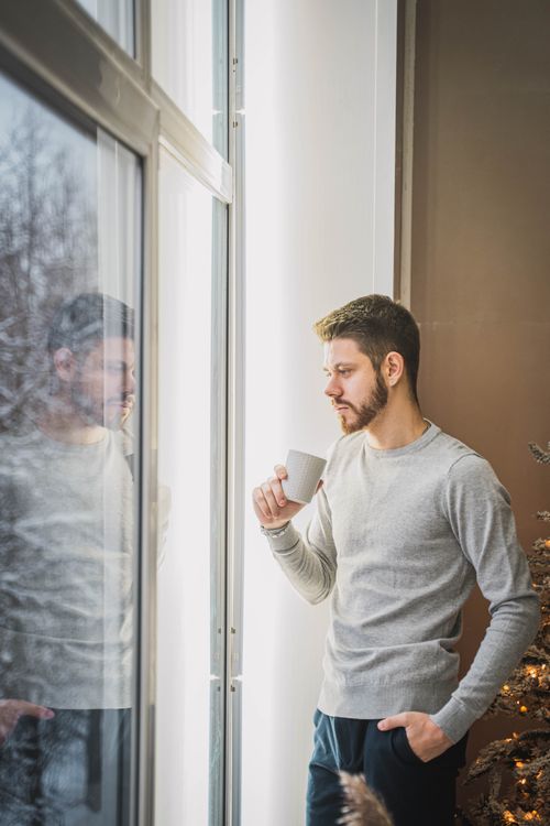Free A man stands by the window, enjoying a cup of coffee, reflecting and relaxing indoors. Stock Photo