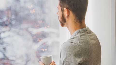 A man stands indoors, holding a coffee cup, staring out a window at a winter landscape.