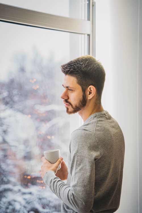Free A young man with a beard gazes thoughtfully out a winter window while holding a cup. Stock Photo